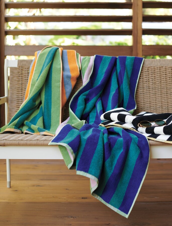 A vibrant Dusen Dusen striped beach towel in the Chincoteague pattern, spread out on sandy beach. The towel features bold, colorful stripes in various widths, creating a playful and eye-catching design. Made from soft, absorbent fabric, it’s perfect for lounging by the water or drying off after a swim, adding a stylish flair to any beach day.